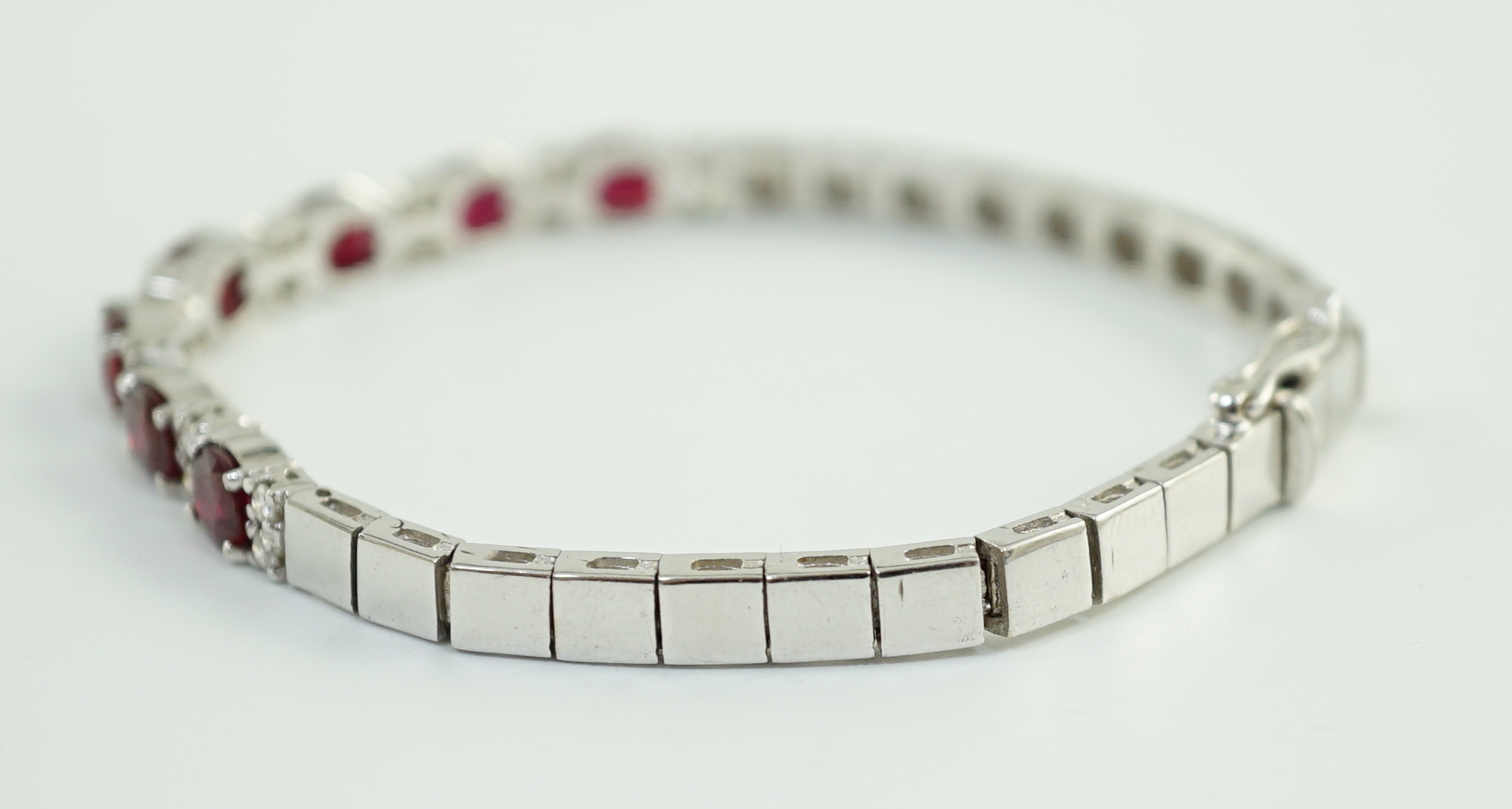 A 750 white gold and seven stone oval cut ruby set line bracelet, set with sixteen stone diamond spacers